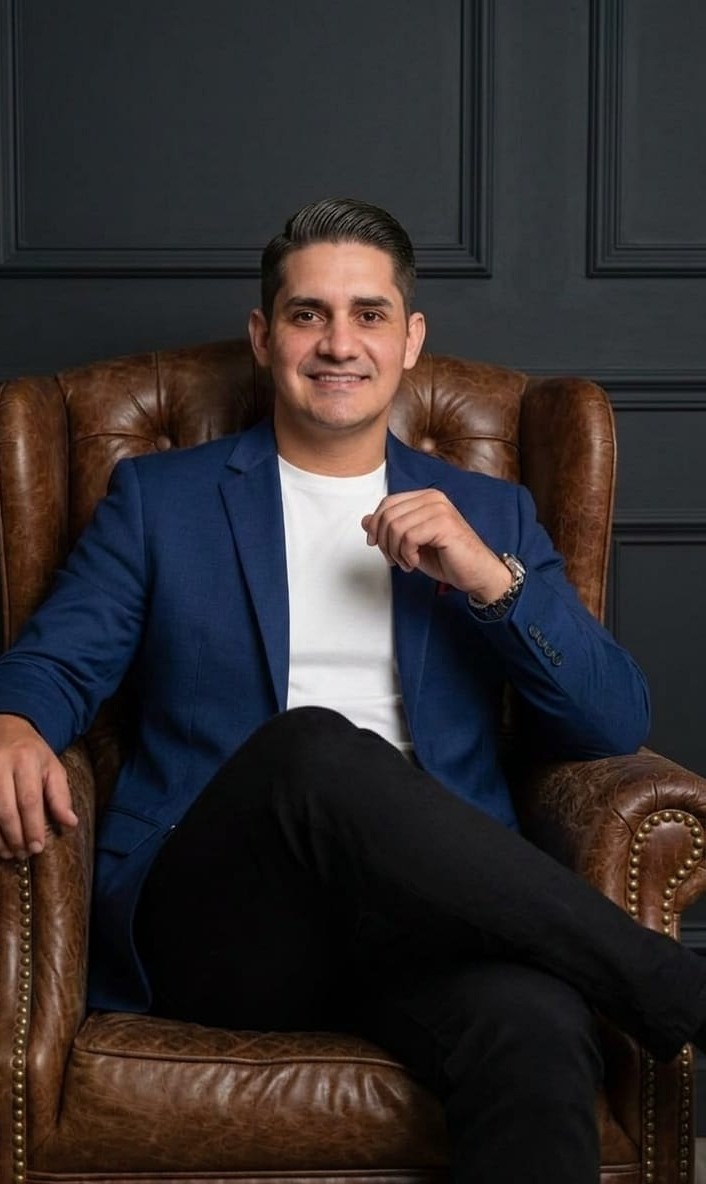 homem sorridente de blazer azul e calça preta sentado numa poltrona de couro marrom contra parede escura